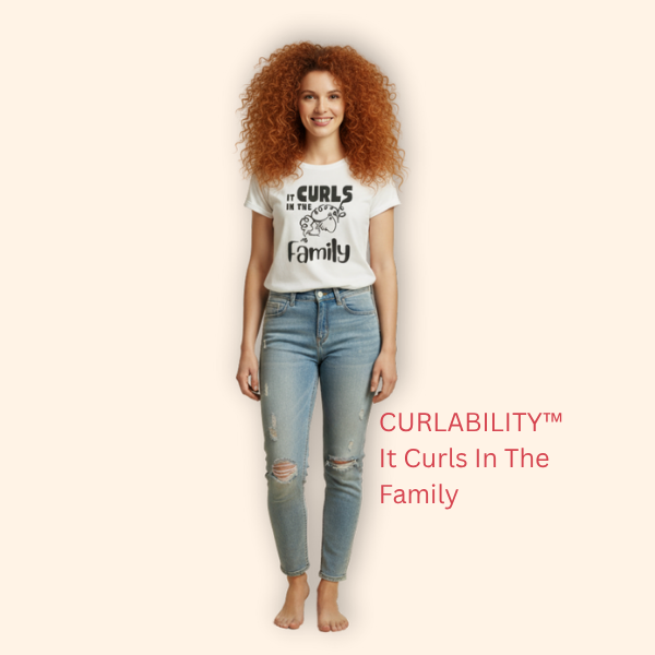 A woman with long curly red hair wearing a white “It Curls In The Family” t-shirt from CURLABILITY™, standing and smiling against a soft beige background.