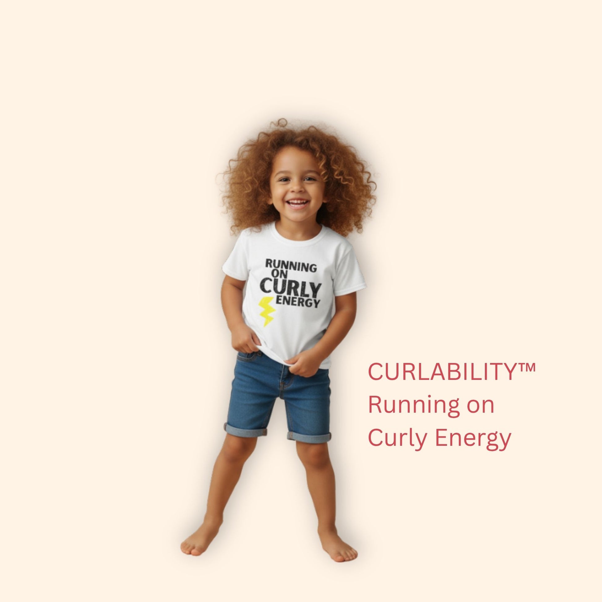 Smiling curly-haired girl standing in a studio wearing a white “Running on Curly Energy” t-shirt and denim shorts.
