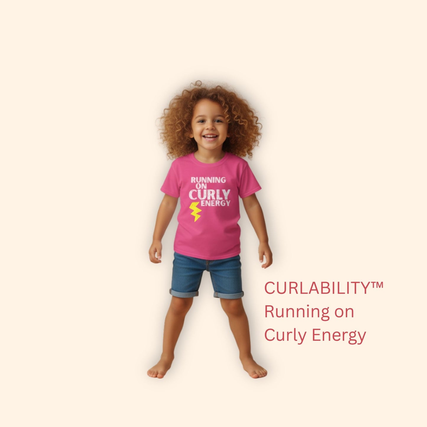 Smiling curly-haired girl standing in a studio wearing a Heliconia “Running on Curly Energy” t-shirt and denim shorts.