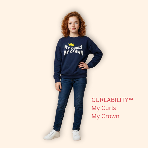 Full-body studio photo of a curly-haired girl wearing a navy “My Curls My Crown” sweatshirt, standing confidently against a soft beige background. Kids’ curly-hair empowerment apparel by Curlability.