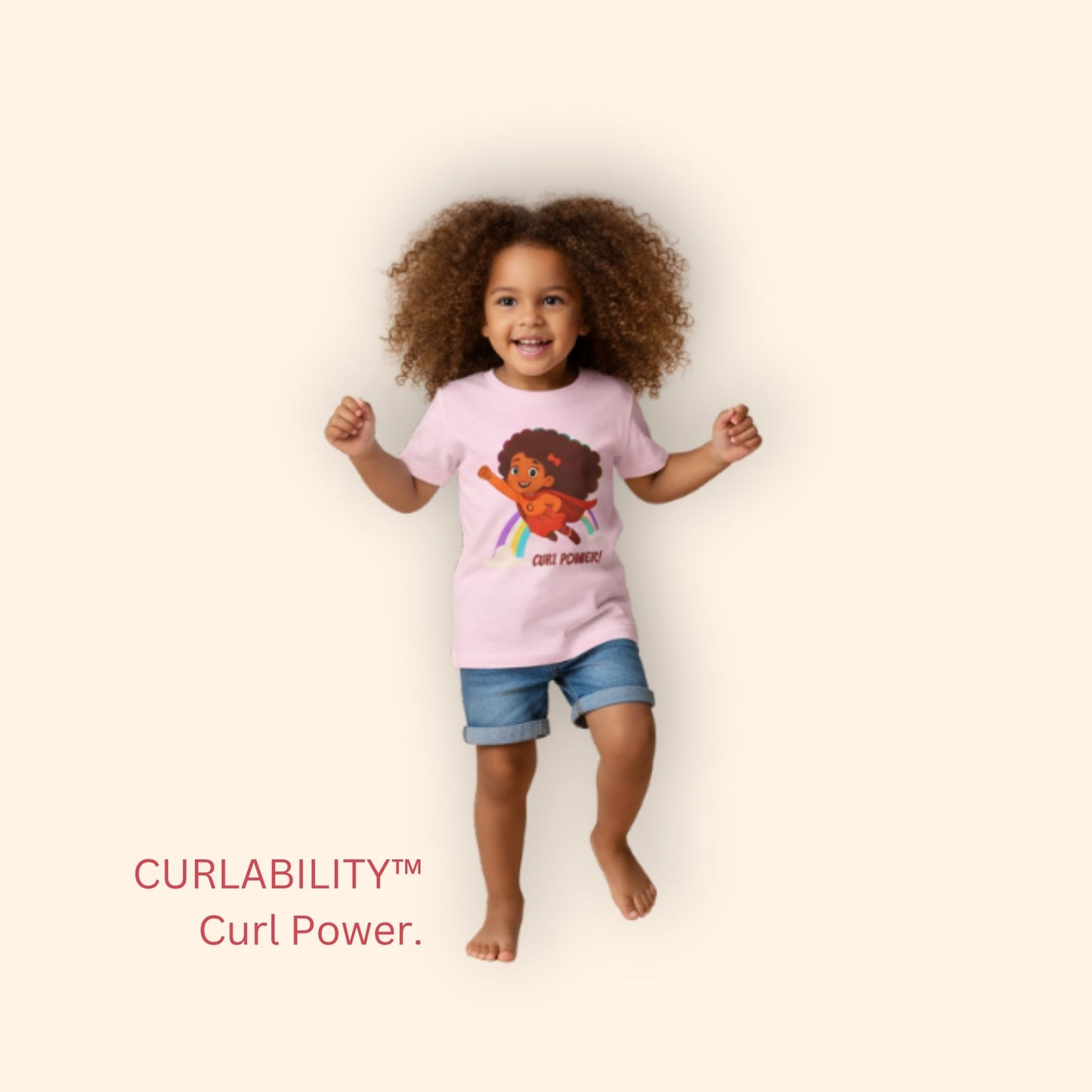 Smiling curly-haired girl standing in a studio wearing a light pink “Curl Power!” t-shirt and denim shorts.