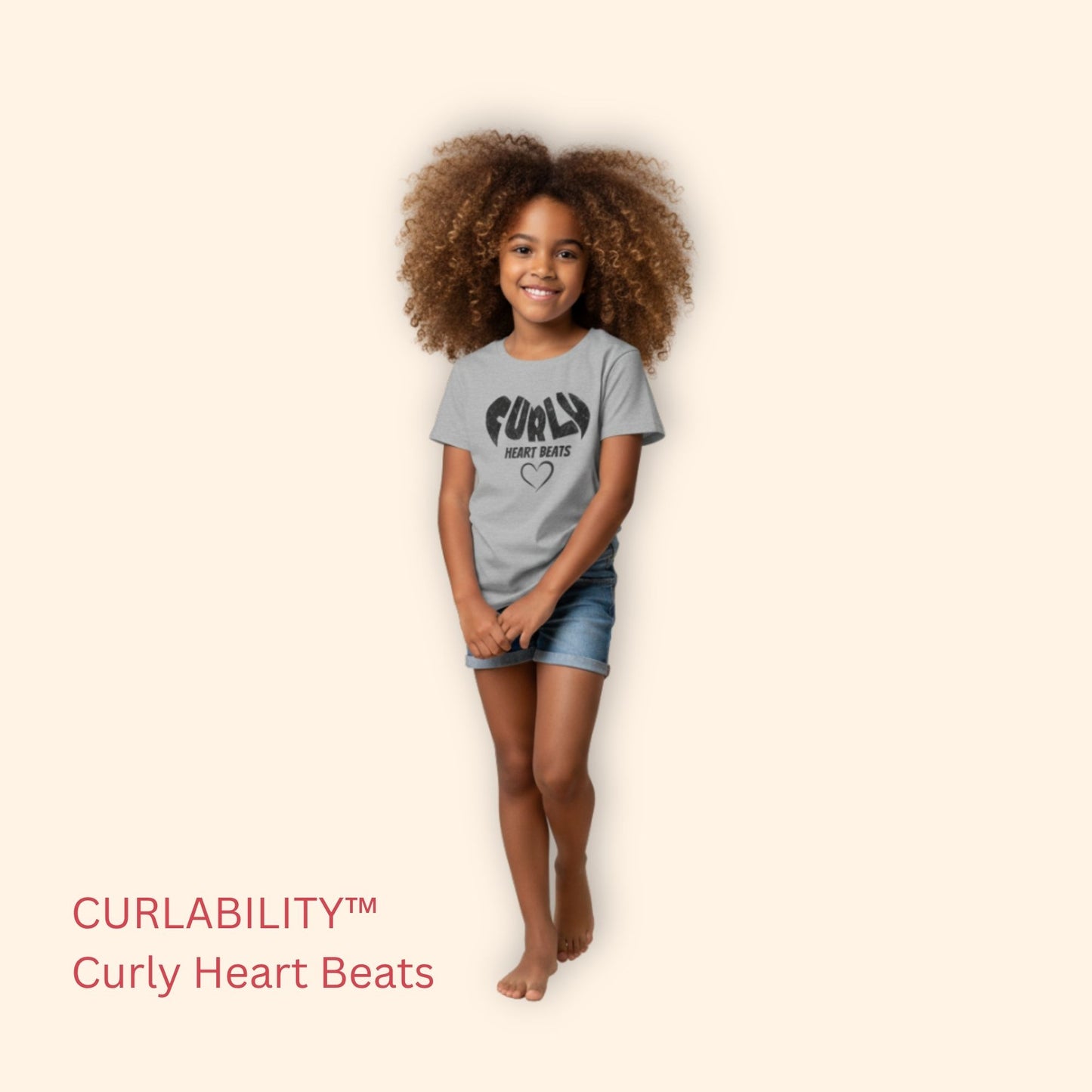 Smiling curly-haired girl standing in a studio wearing a sport grey “Curly Heart Beats” t-shirt and denim shorts.