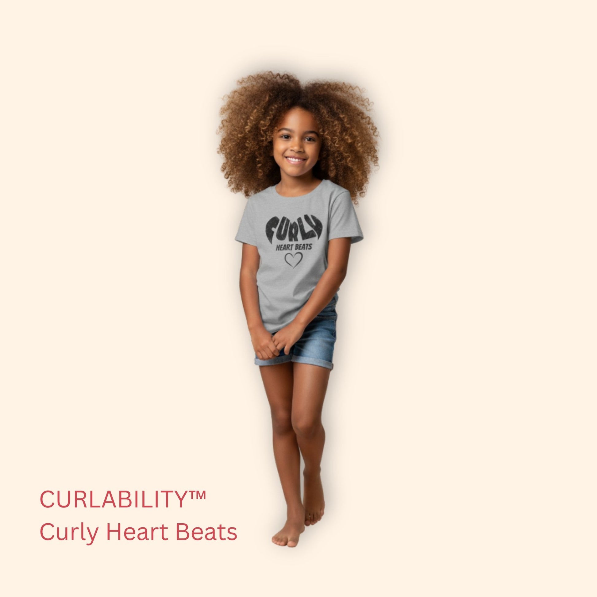 Smiling curly-haired girl standing in a studio wearing a sport grey “Curly Heart Beats” t-shirt and denim shorts.