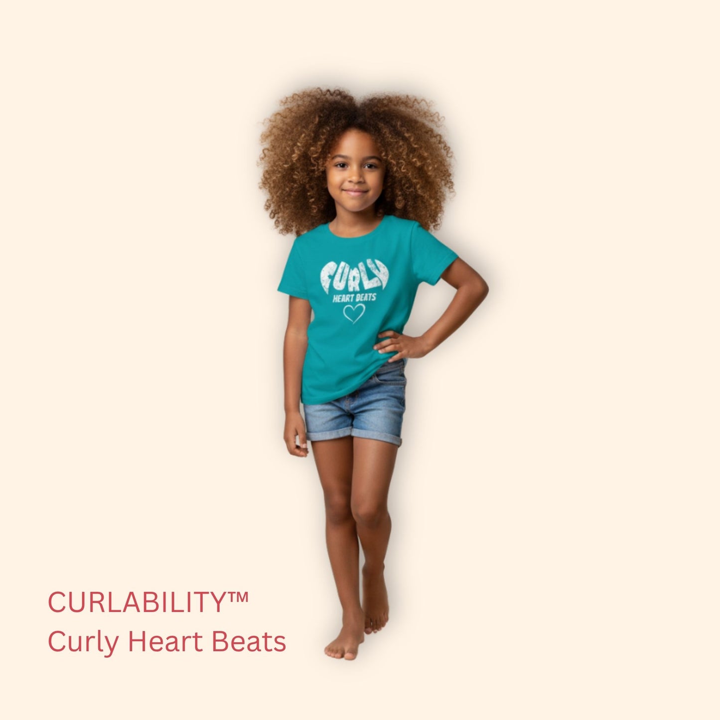 Smiling curly-haired girl standing in a studio wearing a Tropical Blue “Curly Heart Beats” t-shirt and denim shorts.