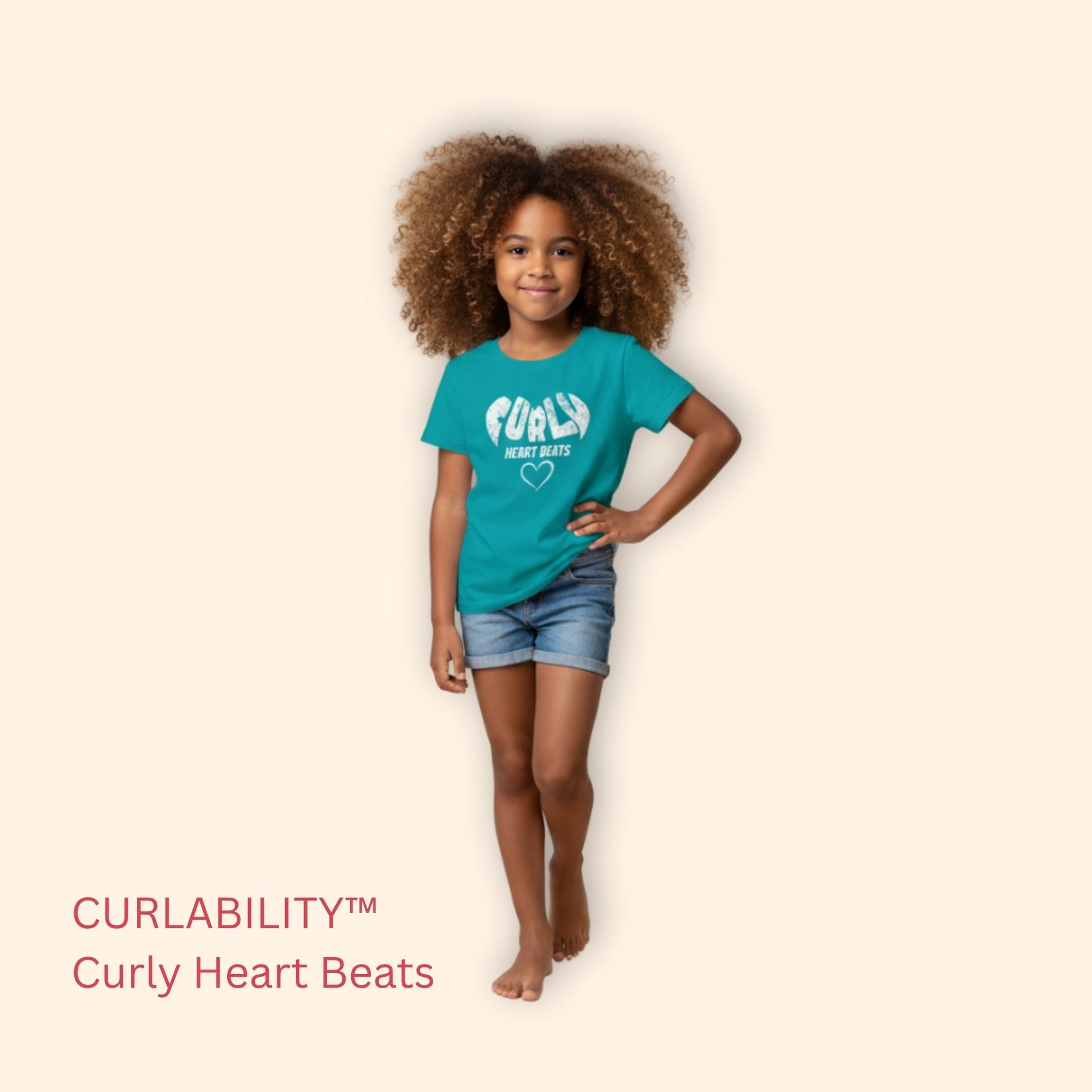 Smiling curly-haired girl standing in a studio wearing a Tropical Blue “Curly Heart Beats” t-shirt and denim shorts.