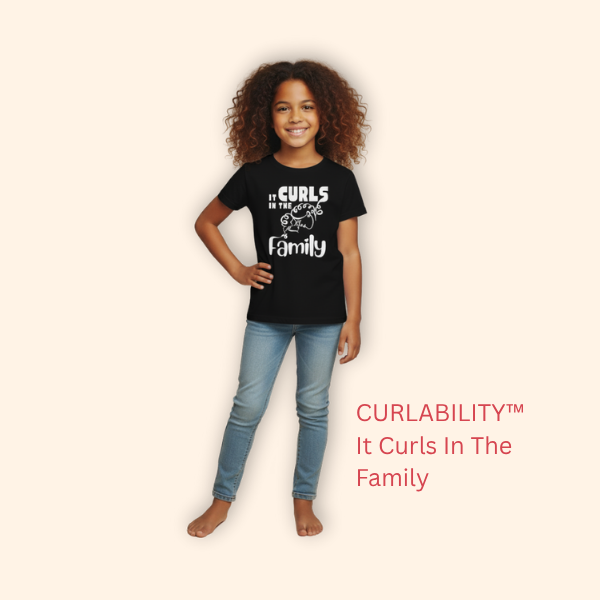Smiling curly-haired girl standing in a studio wearing a black “It Curls in the Family” t-shirt and light blue jeans.