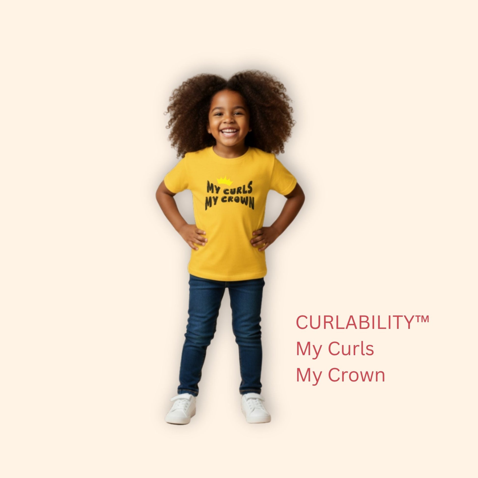 Smiling curly-haired girl standing in a studio wearing a yellow “My Curls My Crown” t-shirt and jeans.