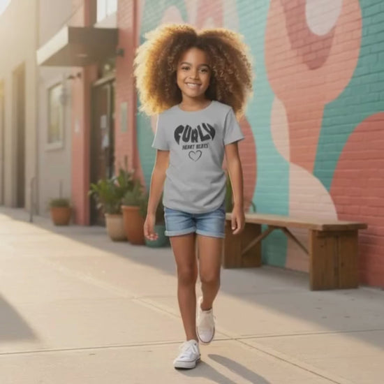 Smiling curly-haired girl walking outdoors wearing a sport grey “Curly Heart Beats” t-shirt and denim shorts.
