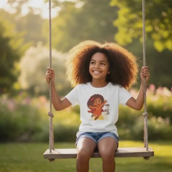 Smiling curly-haired girl sitting on a swing in a sunlit park wearing a white “Curl Up. Rise Up.” t-shirt.
