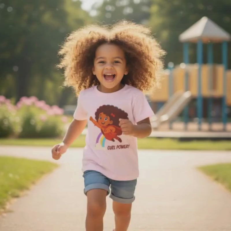 Smiling curly-haired girl running in a park wearing a light pink “Curl Power!” t-shirt.