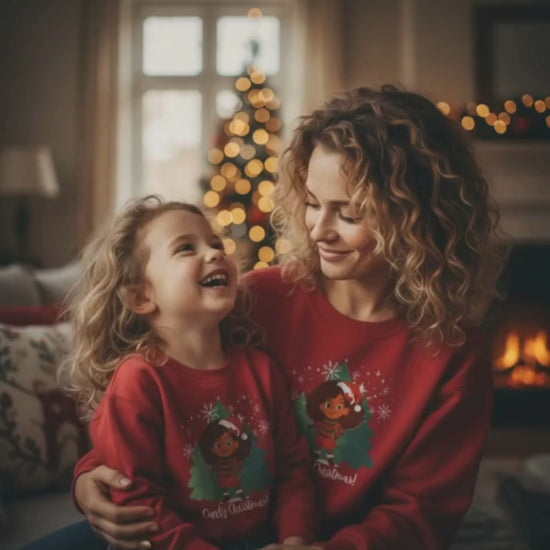 Mother and daughter with curly hair wearing matching red “Curly Christmas” sweatshirts, smiling together in a cozy holiday living room with a Christmas tree glowing in the background.