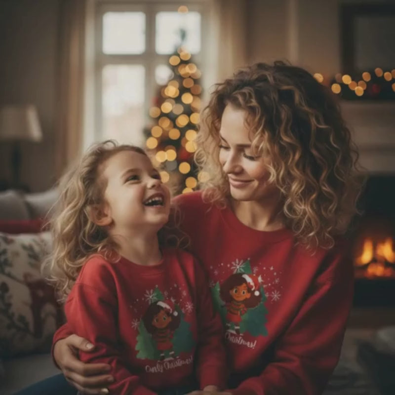 Mother and daughter with curly hair wearing matching red “Curly Christmas” sweatshirts, smiling together in a cozy holiday living room with a Christmas tree glowing in the background.