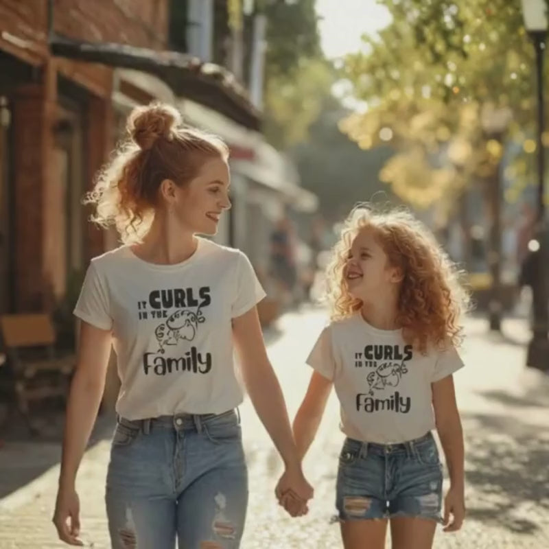 Mother and daughter with curly hair walking outdoors together wearing matching “It Curls in the Family” t-shirts.