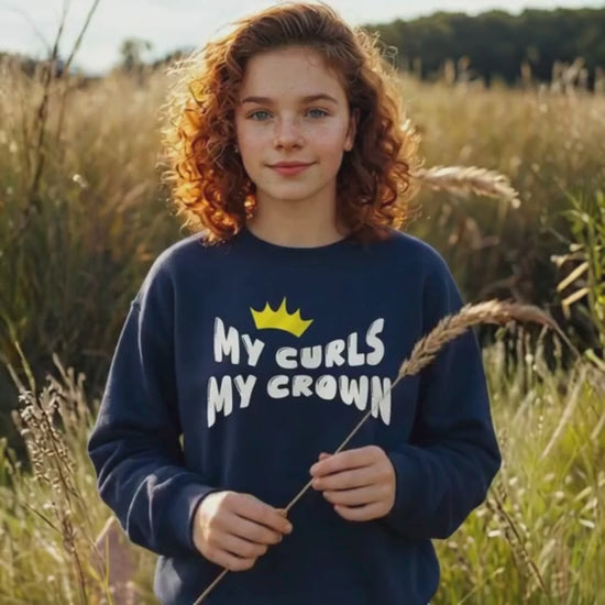 Video of a curly-haired teen girl standing outdoors in a sunlit field, wearing a navy “My Curls My Crown” sweatshirt with a yellow crown graphic. She smiles softly and gently holds a piece of tall grass, highlighting natural curls and empowerment-themed apparel.