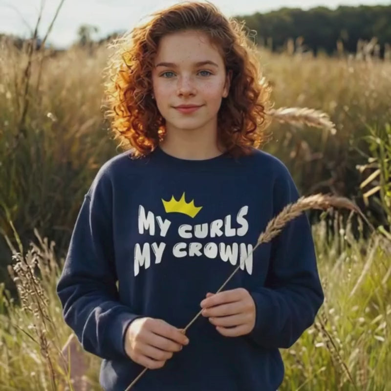 Video of a curly-haired teen girl standing outdoors in a sunlit field, wearing a navy “My Curls My Crown” sweatshirt with a yellow crown graphic. She smiles softly and gently holds a piece of tall grass, highlighting natural curls and empowerment-themed apparel.