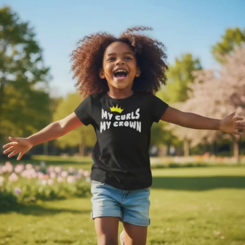 Smiling curly-haired girl running in a park with arms open, wearing a black “My Curls My Crown” t-shirt and denim shorts.