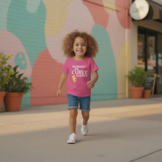 Smiling curly-haired girl walking outdoors wearing a Heliconia “Running on Curly Energy” t-shirt and denim shorts.