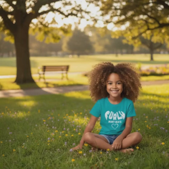 Smiling curly-haired girl sitting on the grass in a park wearing a Tropical Blue “Curly Heart Beats” t-shirt.