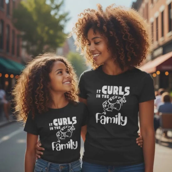 Mother and daughter with curly hair smiling at each other while walking outdoors, both wearing black “It Curls in the Family” t-shirts.