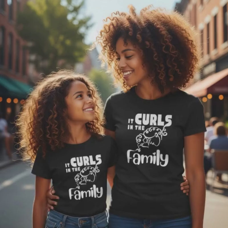 Mother and daughter with curly hair smiling at each other while walking outdoors, both wearing black “It Curls in the Family” t-shirts.