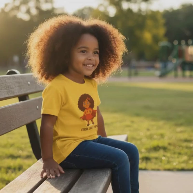 Smiling curly-haired girl sitting on a park bench wearing a yellow “Strong. Curly. Proud.” t-shirt.
