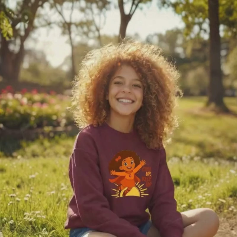 Video of a smiling curly-haired girl outdoors wearing a maroon “Curl Up. Rise Up.” kids’ sweatshirt, sitting in a sunny park.