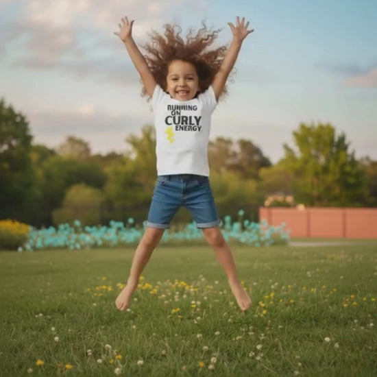 Smiling curly-haired girl jumping in a grassy park wearing a white “Running on Curly Energy” t-shirt and denim shorts.