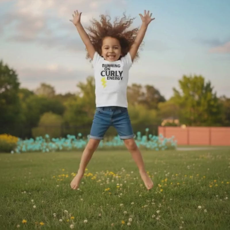 Smiling curly-haired girl jumping in a grassy park wearing a white “Running on Curly Energy” t-shirt and denim shorts.